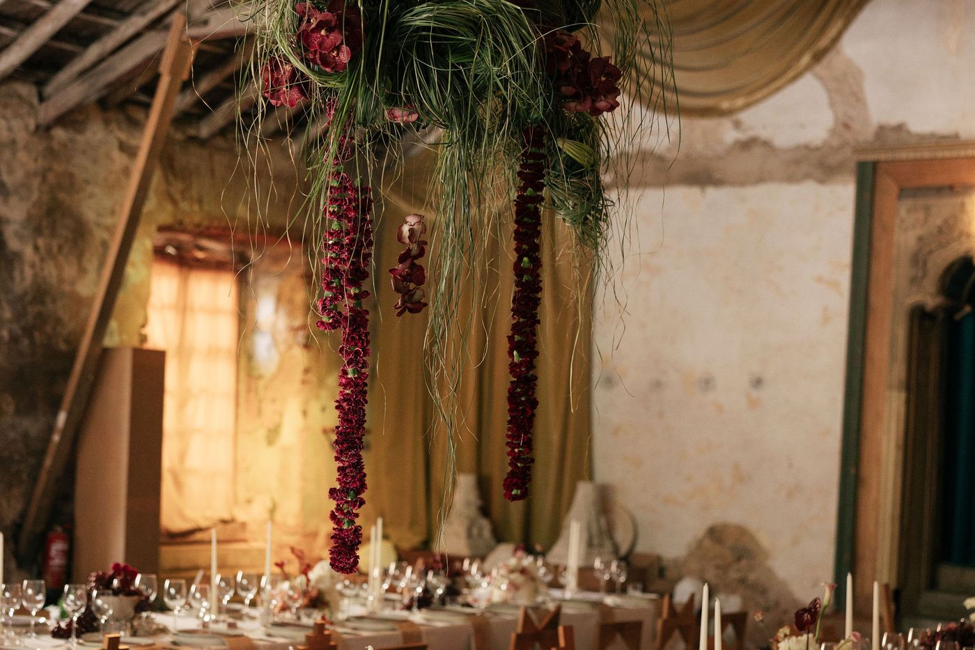 Burgundy garlands hanging above long tables in a candlelit ruin at Pal&aacute;cio do Grilo