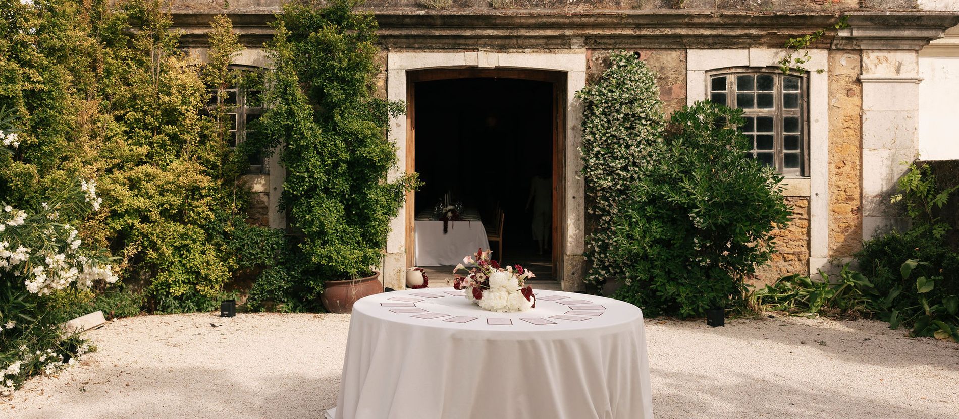 A round seating table dressed in white linen in a gravel courtyard before an ivy-covered stone building